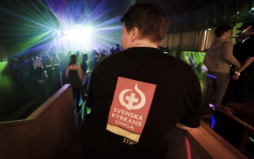 En ungdom har en tröja med texten Svenska kyrkans unga på ryggen. I bakgrunden syns ett fullt dansgolv med discoljus.