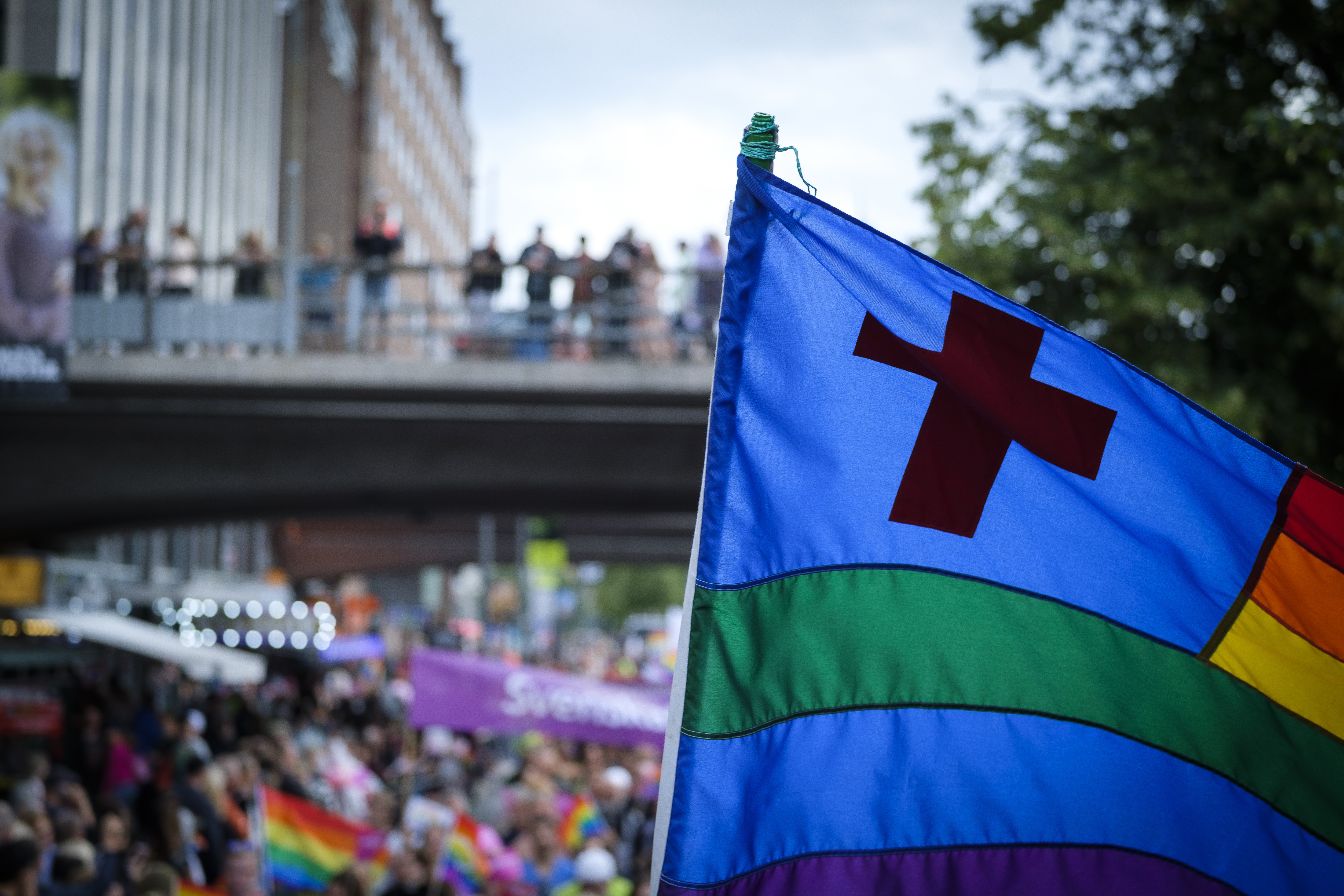 En prideflagga med ett kors. I bakgrunden en folkmassa med andra prideflaggor.