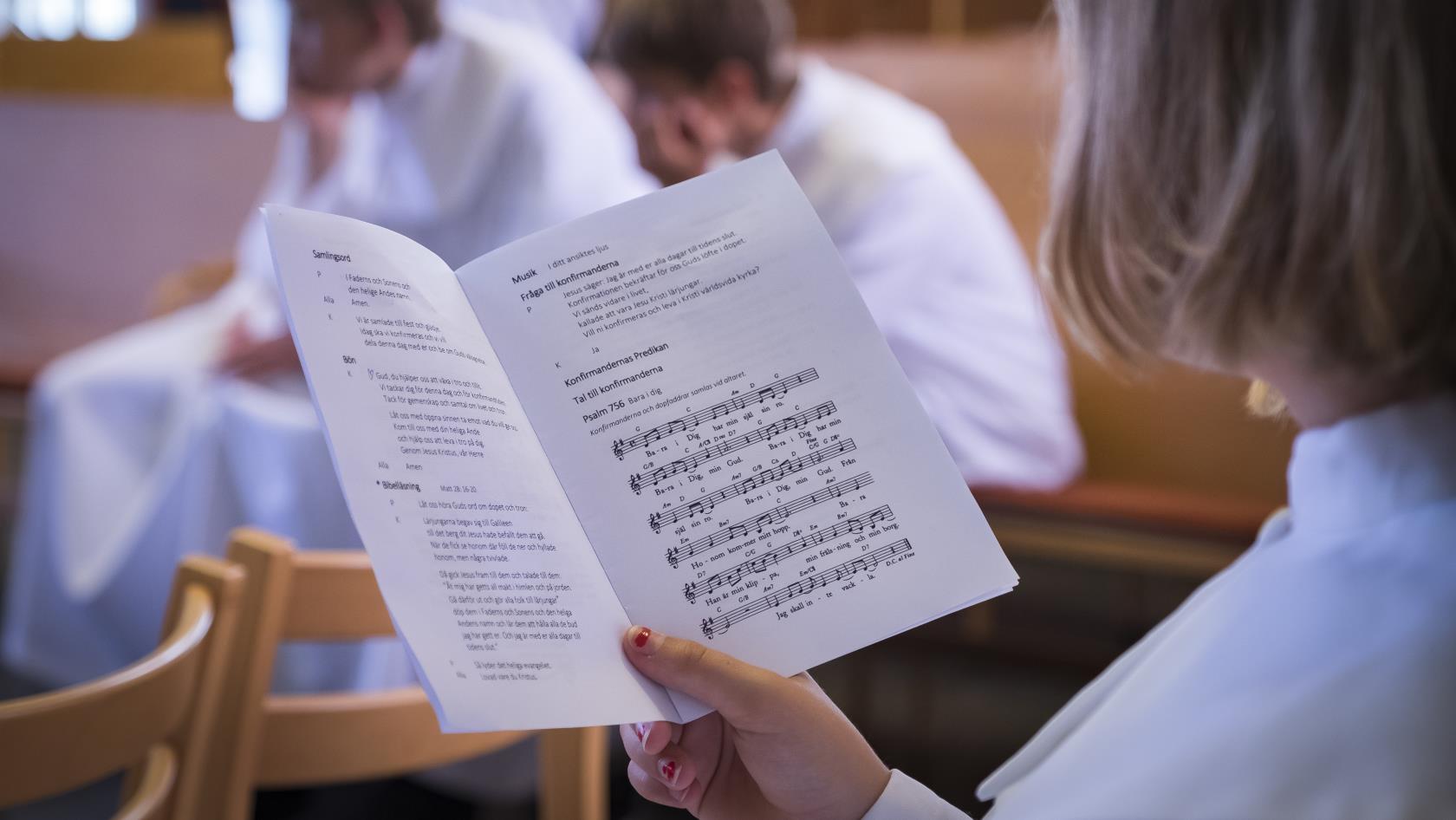 En konfirmand läser i ett sånghäfte. Fler konfirmander sitter i bakgrunden.