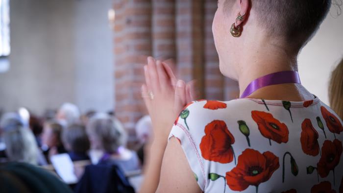 Närbild på en kvinna som står upp med knäppta händer i en kyrka. Fyllda kyrkbänkar syns suddigt i bakgrunden.