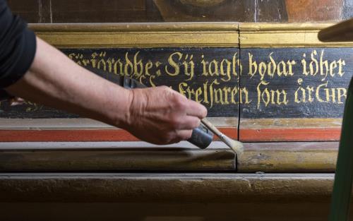 En person dammsuger försiktigt, med hjälp av en pensel, en gammal trähylla i en kyrka.