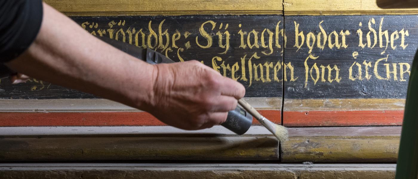 En person dammsuger försiktigt, med hjälp av en pensel, en gammal trähylla i en kyrka.