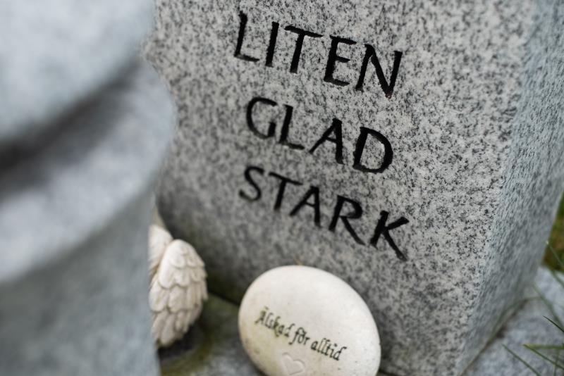 Orden Liten, glad, stark är inristade på en gravsten. Framför ligger en liten vit sten med texten Älskad för alltid.