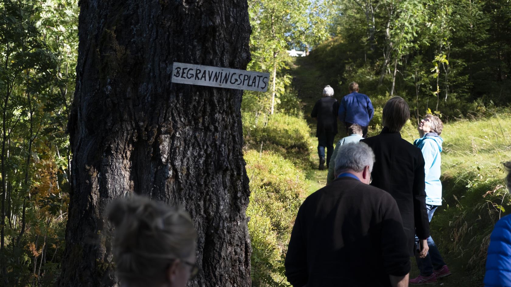 Människor går på en stig förbi ett träd med en skylt med texten Begravningsplats.