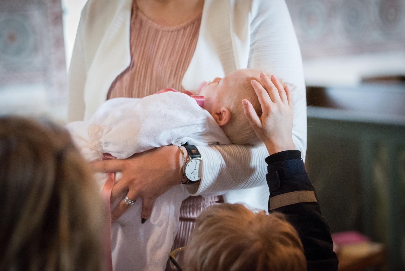 En bebis i dopklänning sover i någons famn. Ett barn klappar på bebisens huvud.