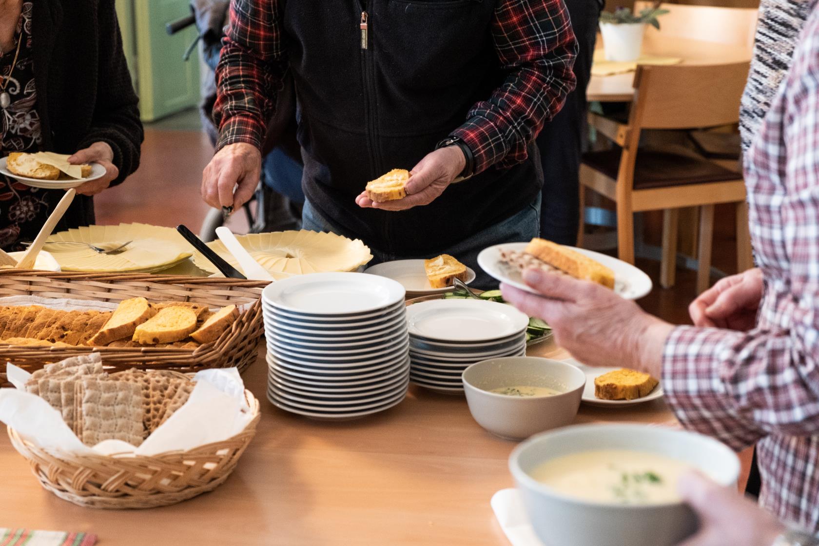 Runt ett fikabord står några personer och brer sina smörgåsar.