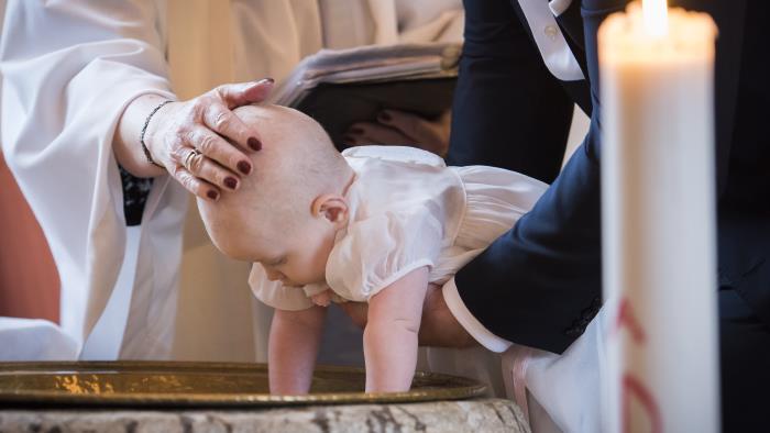 Någon sänker en bebis ner i dopfunten medan prästen håller sin hand på barnets huvud.