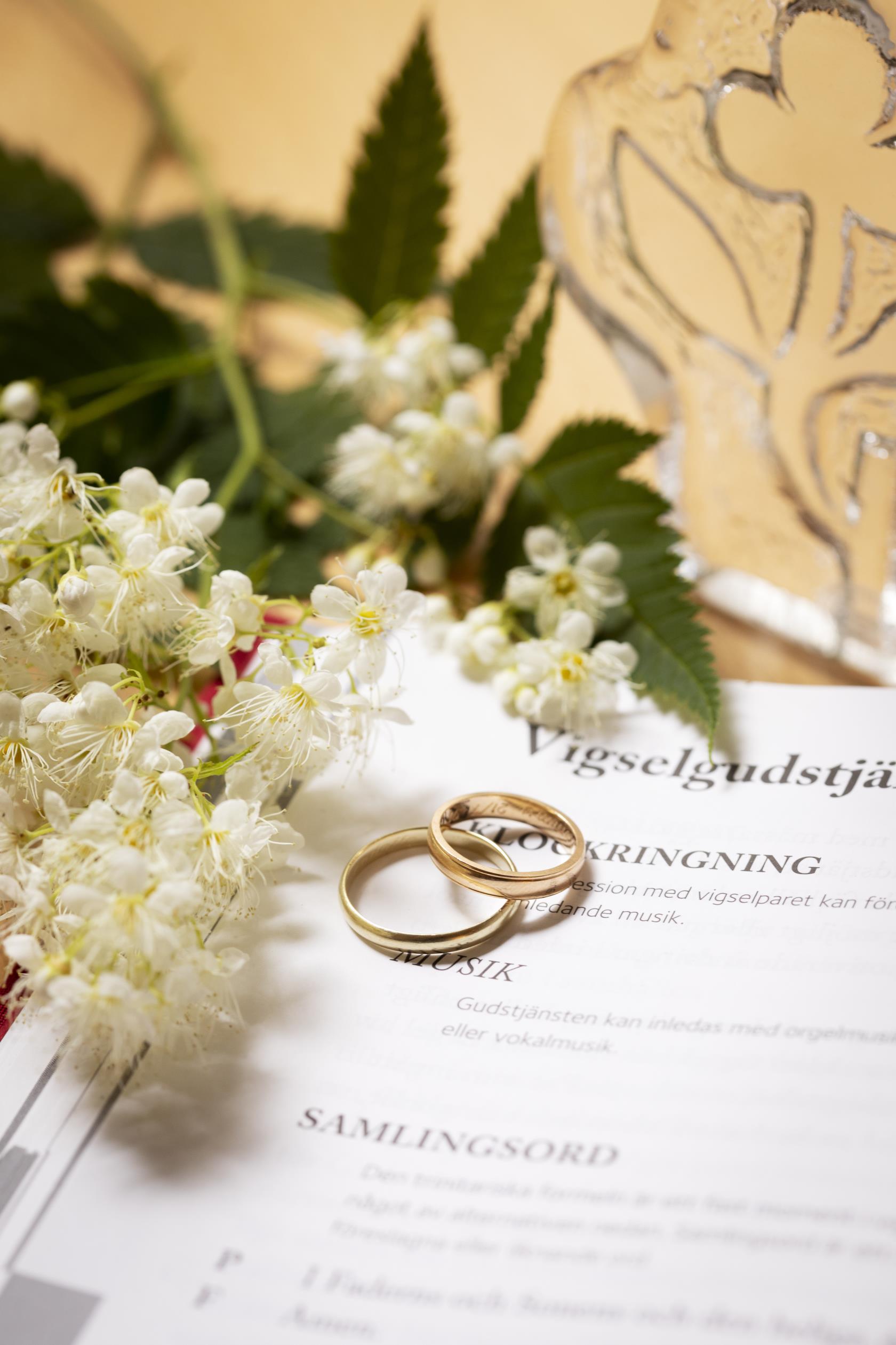 Närbild på två vigselringar som tillsammans med en blomkvist ligger på en uppslagen bok med rubriken Vigselgudstjänst.