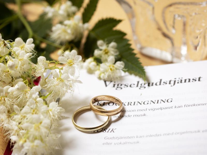 Närbild på två vigselringar som tillsammans med en blomkvist ligger på en uppslagen bok med rubriken Vigselgudstjänst.