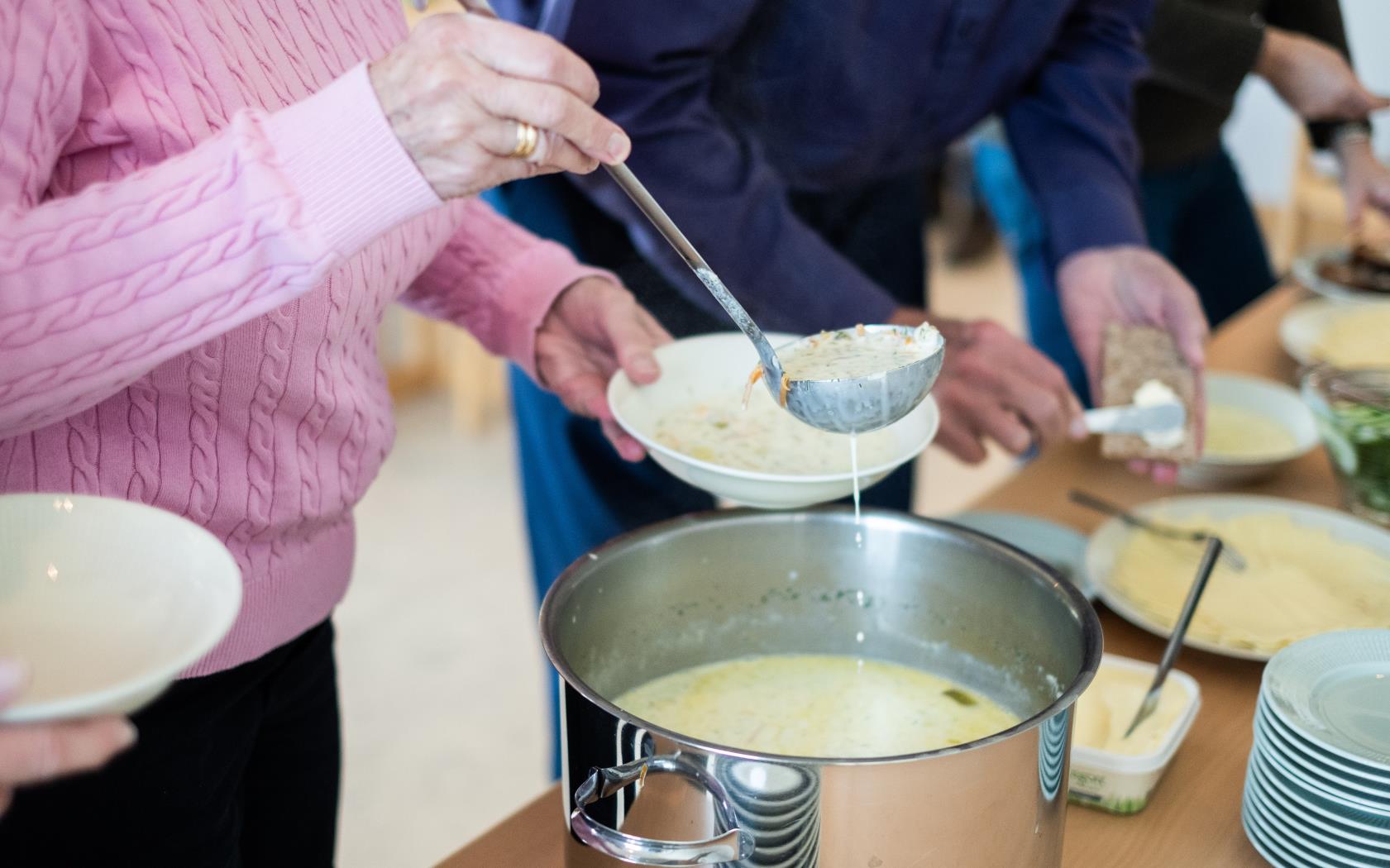 Någon tar en slev soppa ur en kastrull. Bredvid står någon och brer en knäckemacka.