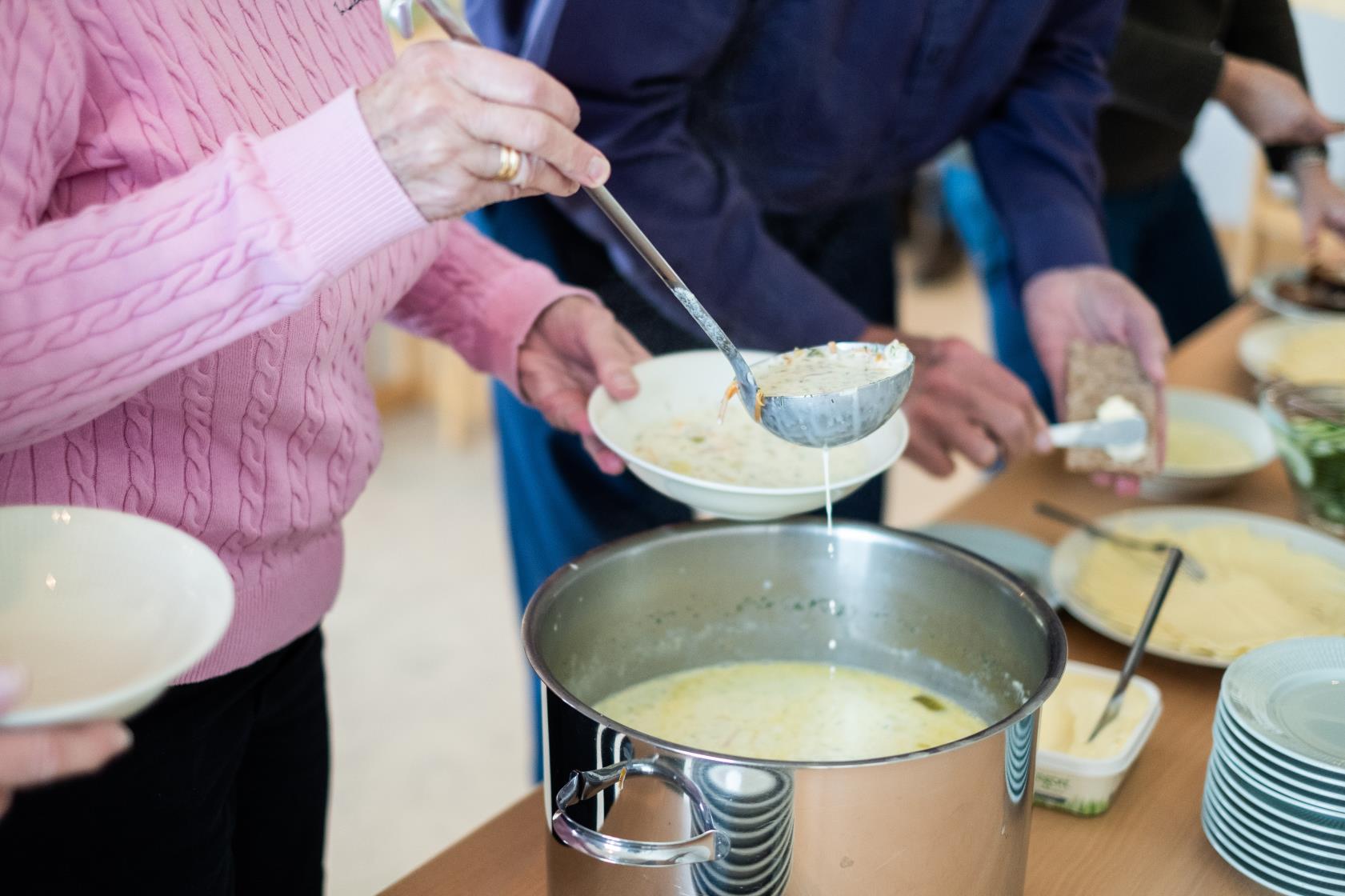 Någon tar en slev soppa ur en kastrull. Bredvid står någon och brer en knäckemacka.