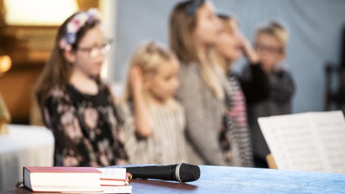 En psalmbok och en mikrofon ligger framlagda. I bakgrunden syns sjungande barn.