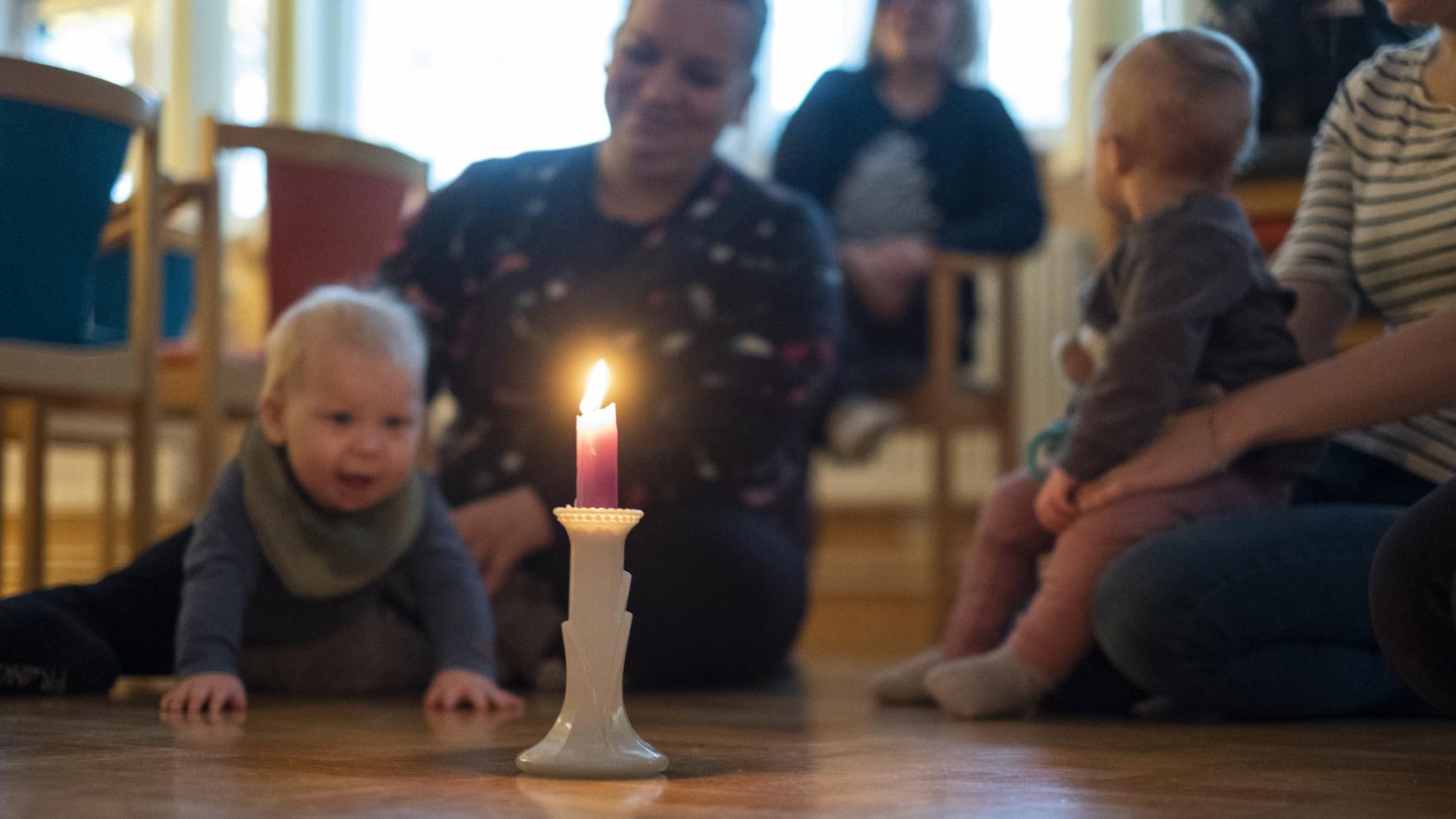 Några mammor sitter i en ring på golvet med sina bebisar. En bebis tittar nyfiket på ett levande ljus som står i mitten.