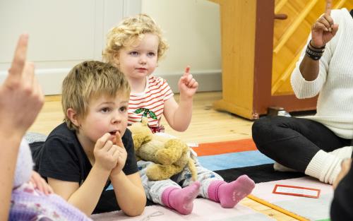 Två barn sitter i ring på golvet och pekar uppåt tillsammans med en ledare.