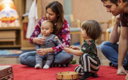 Ett litet barn spelar på en tamburin som ligger på en röd matta. Andra barn och vuxna sitter runt omkring.