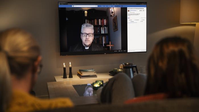 Två personer sitter i en soffa med ryggen mot kameran. På en TV-skärm, med levande ljus uppställda framför, syns en präst.