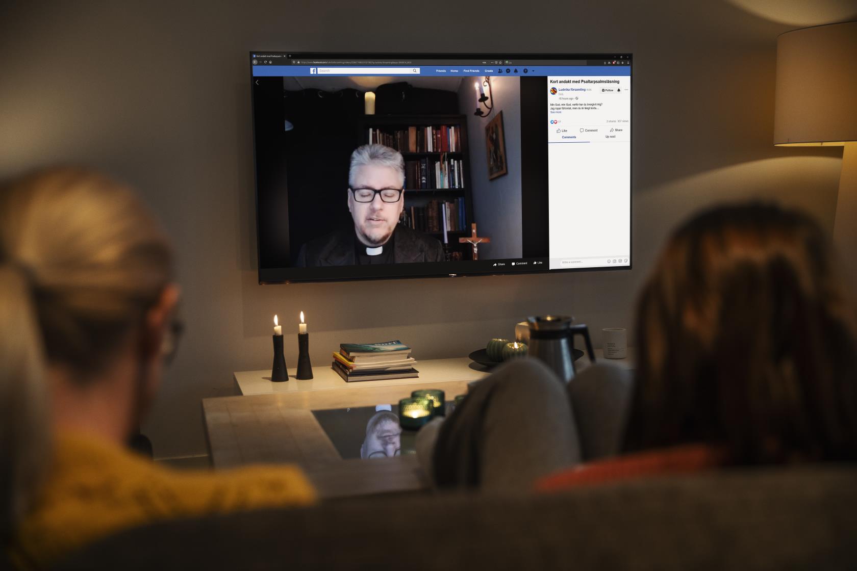 Två personer sitter i en soffa med ryggen mot kameran. På en TV-skärm, med levande ljus uppställda framför, syns en präst.