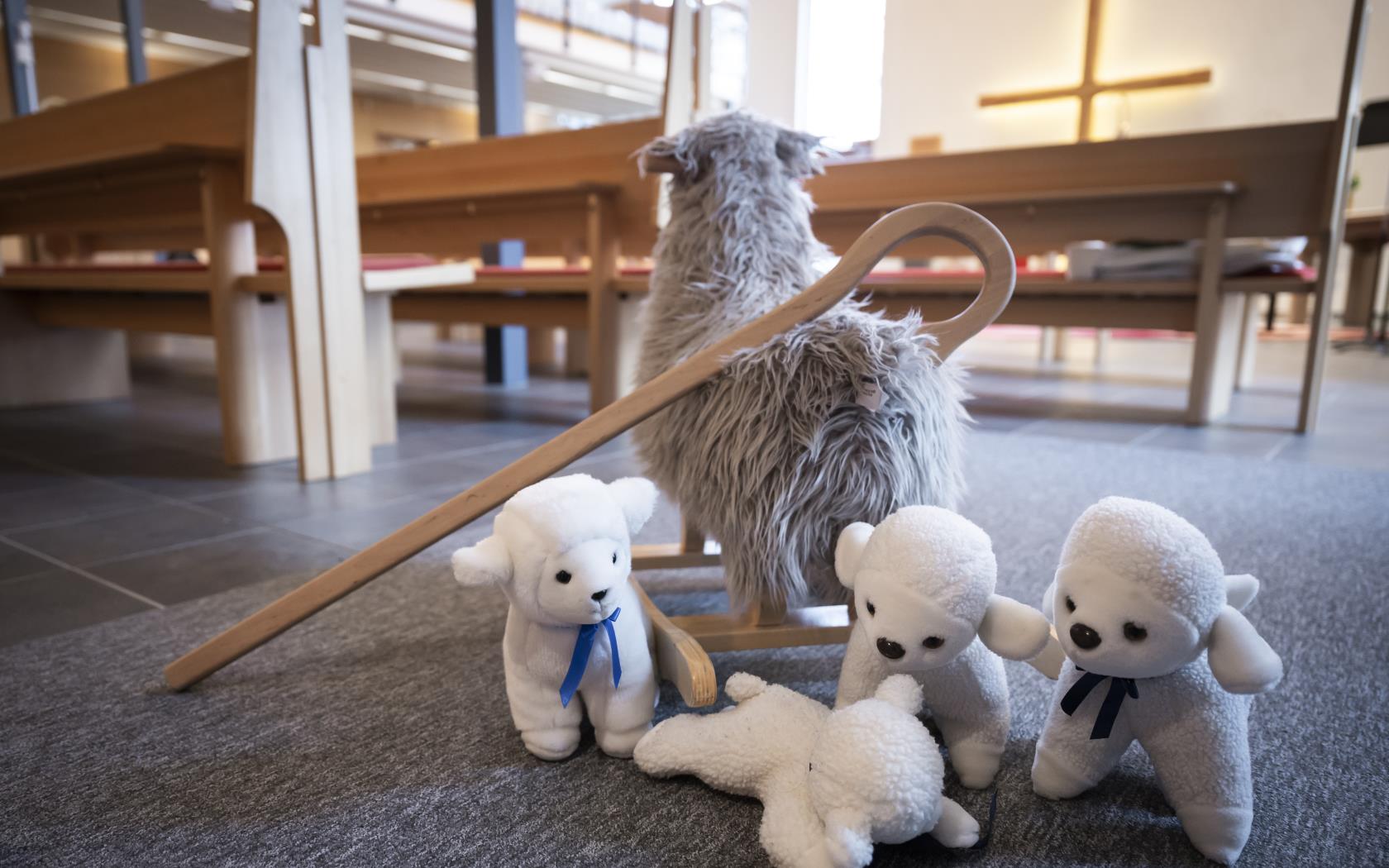 Några lamm- och fårgosedjur och en träkäpp står på golvet i en kyrka.