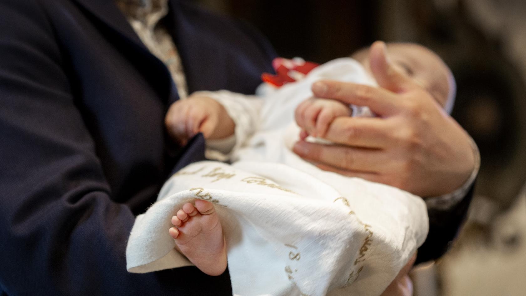 En bebis i dopklänning vilar i någons famn.