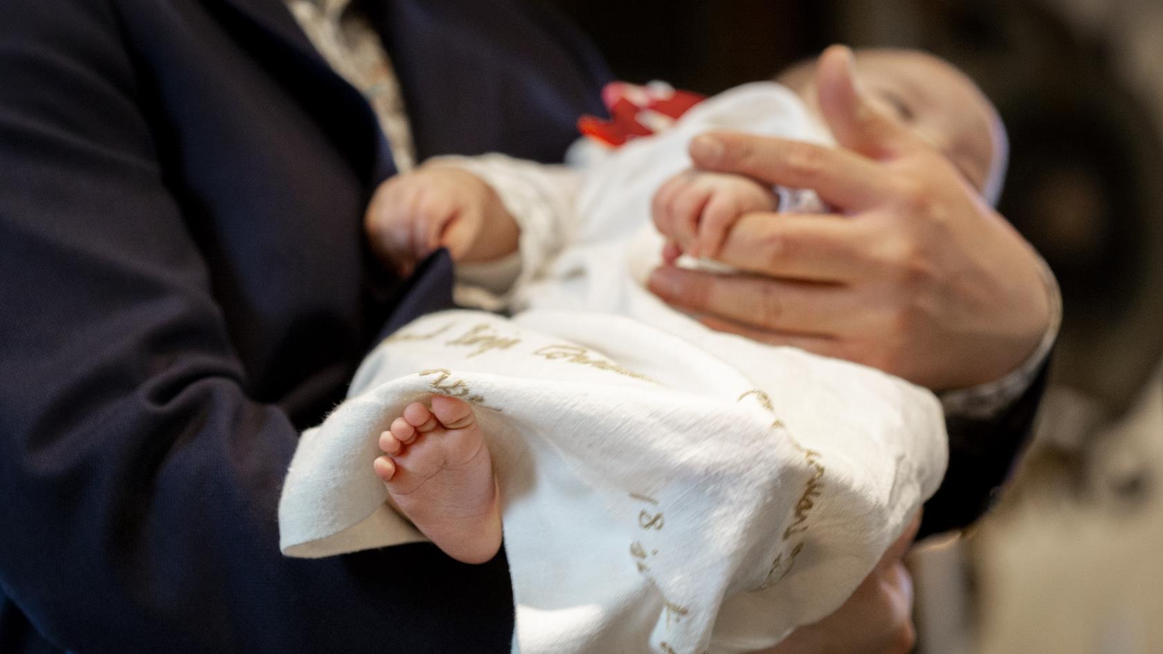 En bebis i dopklänning vilar i någons famn.