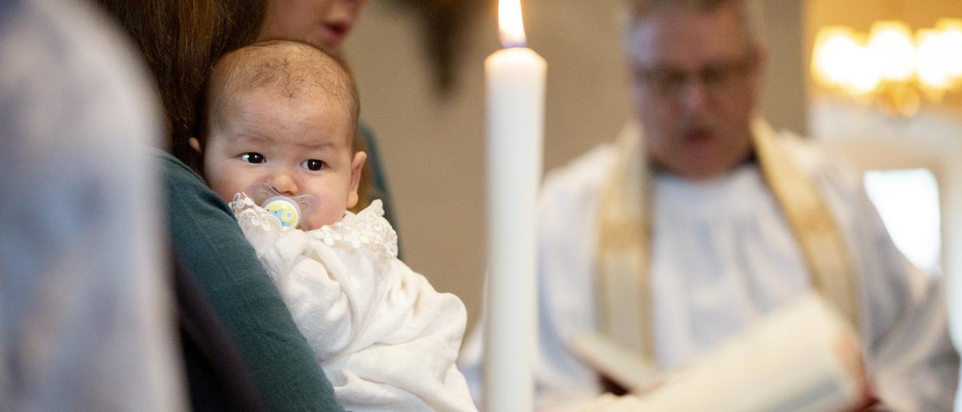 En bebis i dopklänning vilar i någons famn. I bakgrunden står en präst.