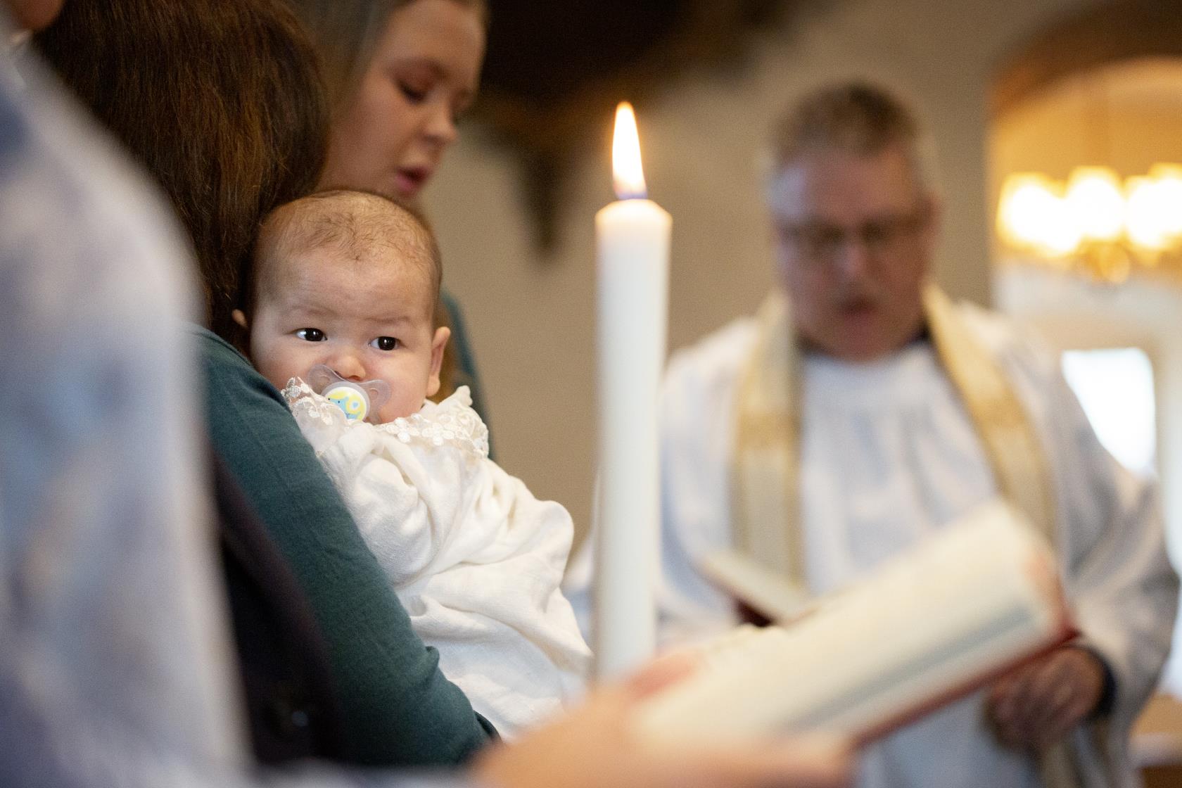 En bebis i dopklänning vilar i någons famn. I bakgrunden står en präst.