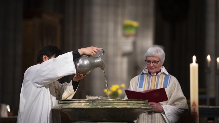 En präst i vit kåpa häller vatten i Uppsala domkyrkas dopfunt. Ärkebiskop Antje Jackelén står bredvid.