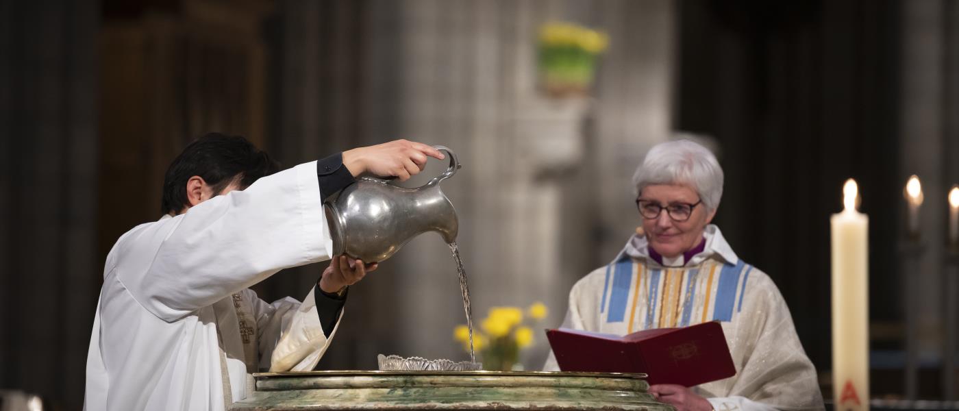 En präst i vit kåpa häller vatten i Uppsala domkyrkas dopfunt. Ärkebiskop Antje Jackelén står bredvid.