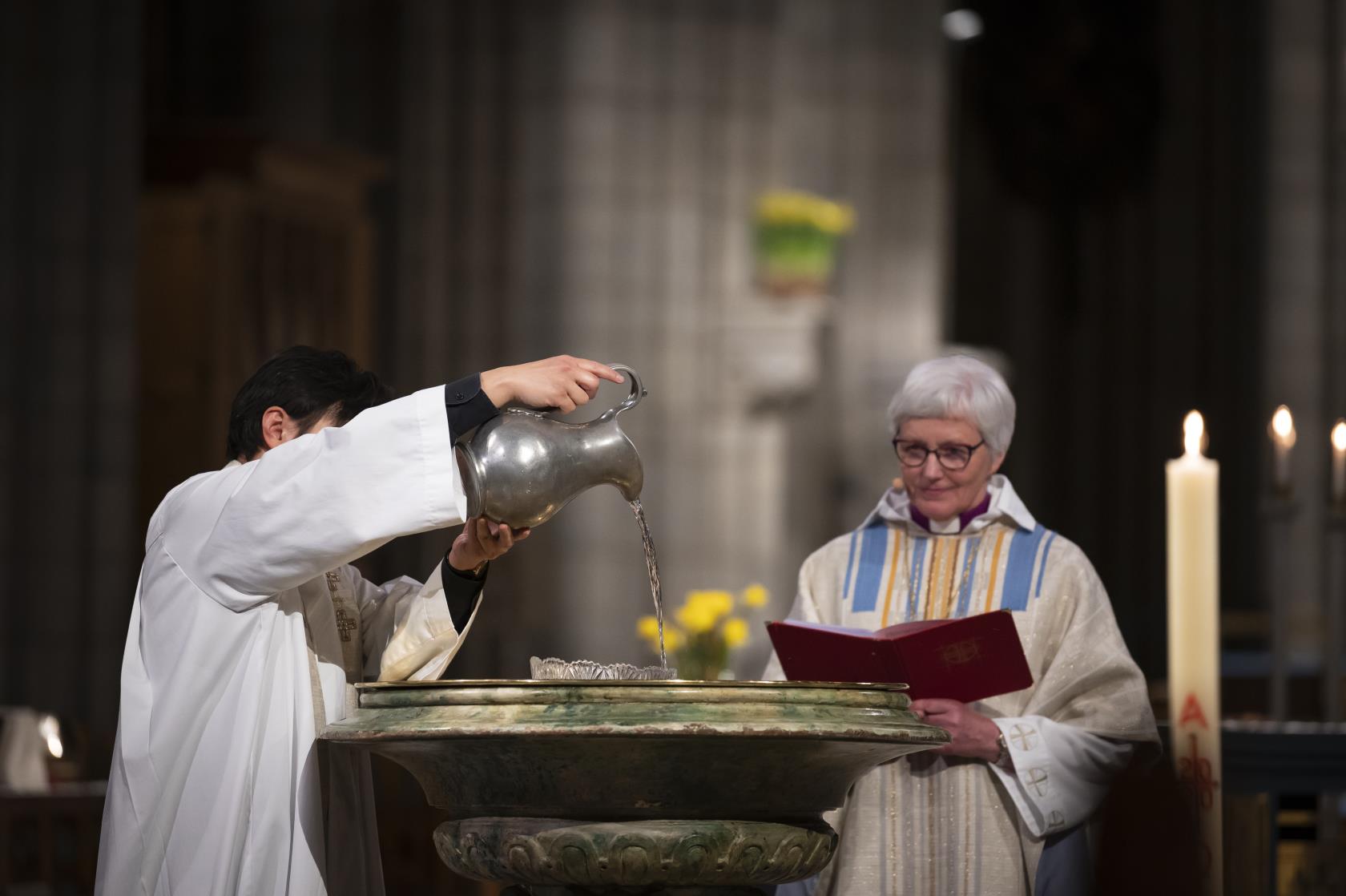 En präst i vit kåpa häller vatten i Uppsala domkyrkas dopfunt. Ärkebiskop Antje Jackelén står bredvid.