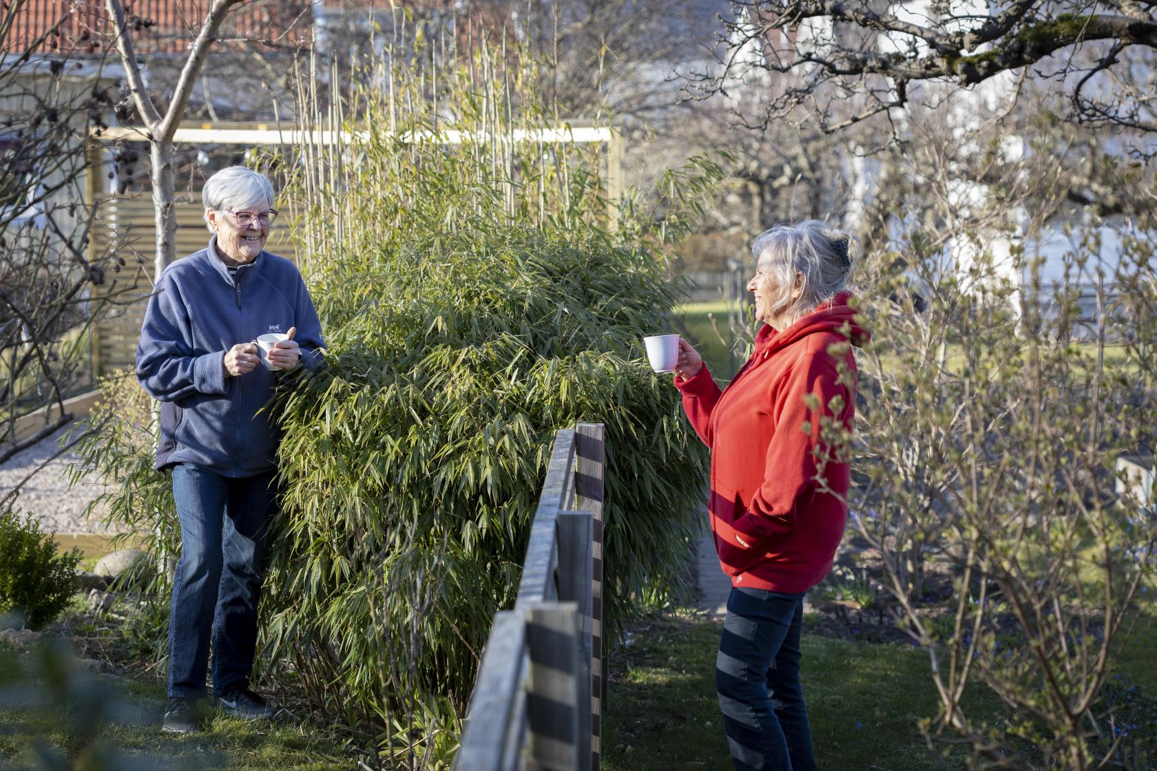 Två grannar samtalar över staketet med var sin kaffekopp i handen.