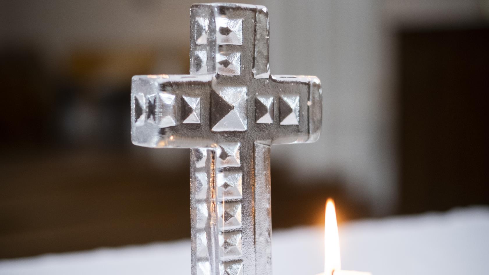 Ett kors av handgjort glas står på ett altare. Framför korset brinner ett ljus på en trefotad ljusstake.