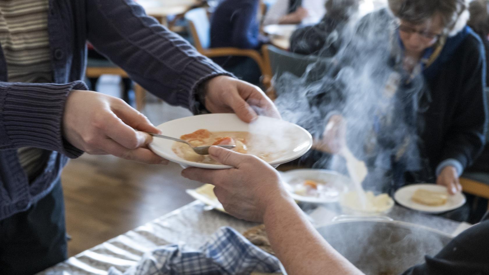 En person serverar soppa och bröd till äldre.