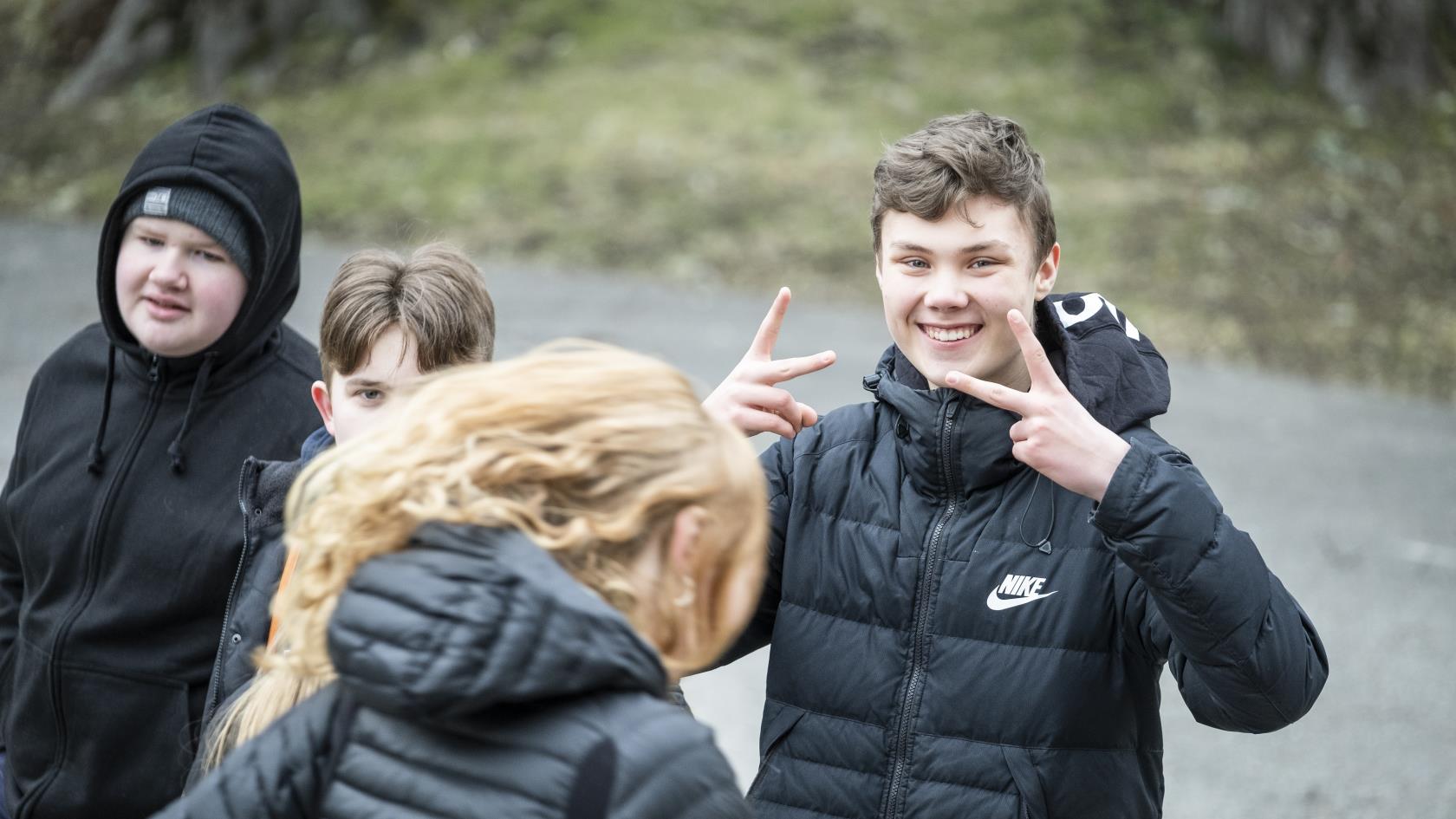 En ung kille ler mot kameran och gör peace-tecken med händerna. Hans kompisar står bredvid.