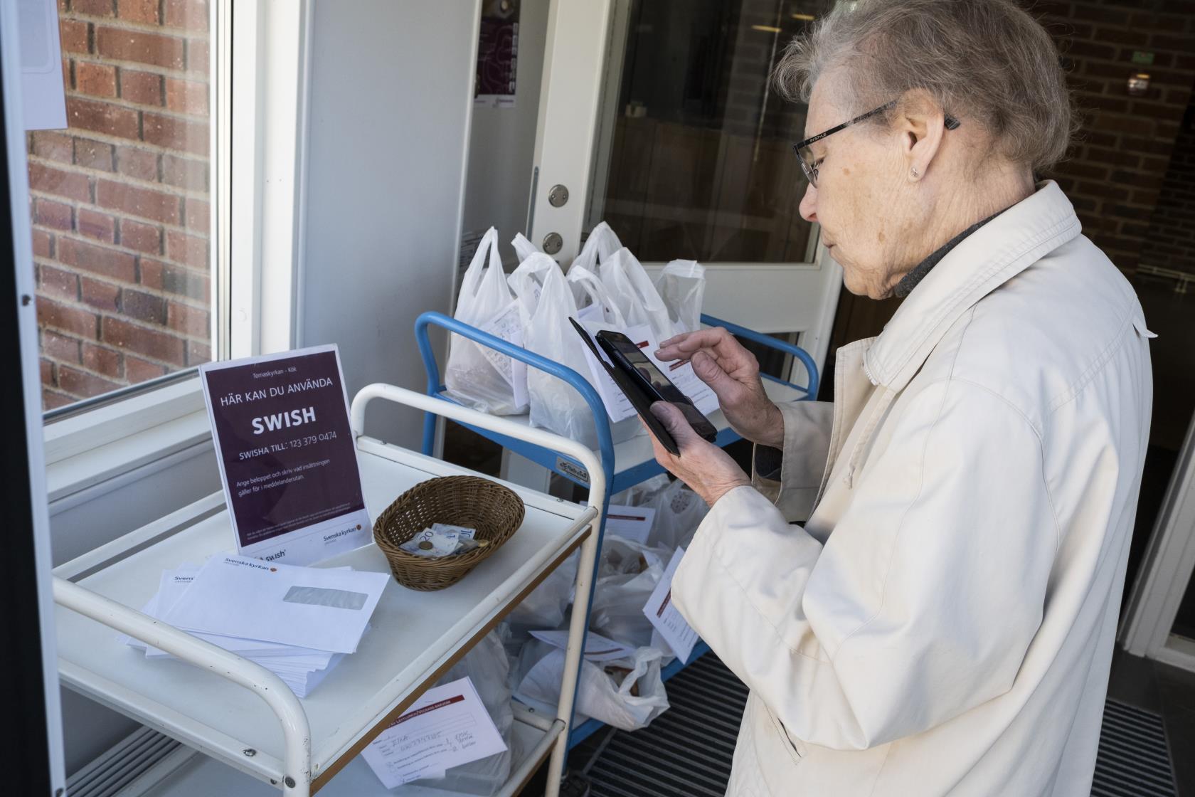 En äldre dam står med mobiltelefonen framför en skylt med texten Här kan du använda Swish.