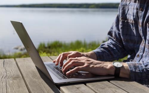 En person sitter vid ett träbord vid en sjö och jobbar på sin laptop.