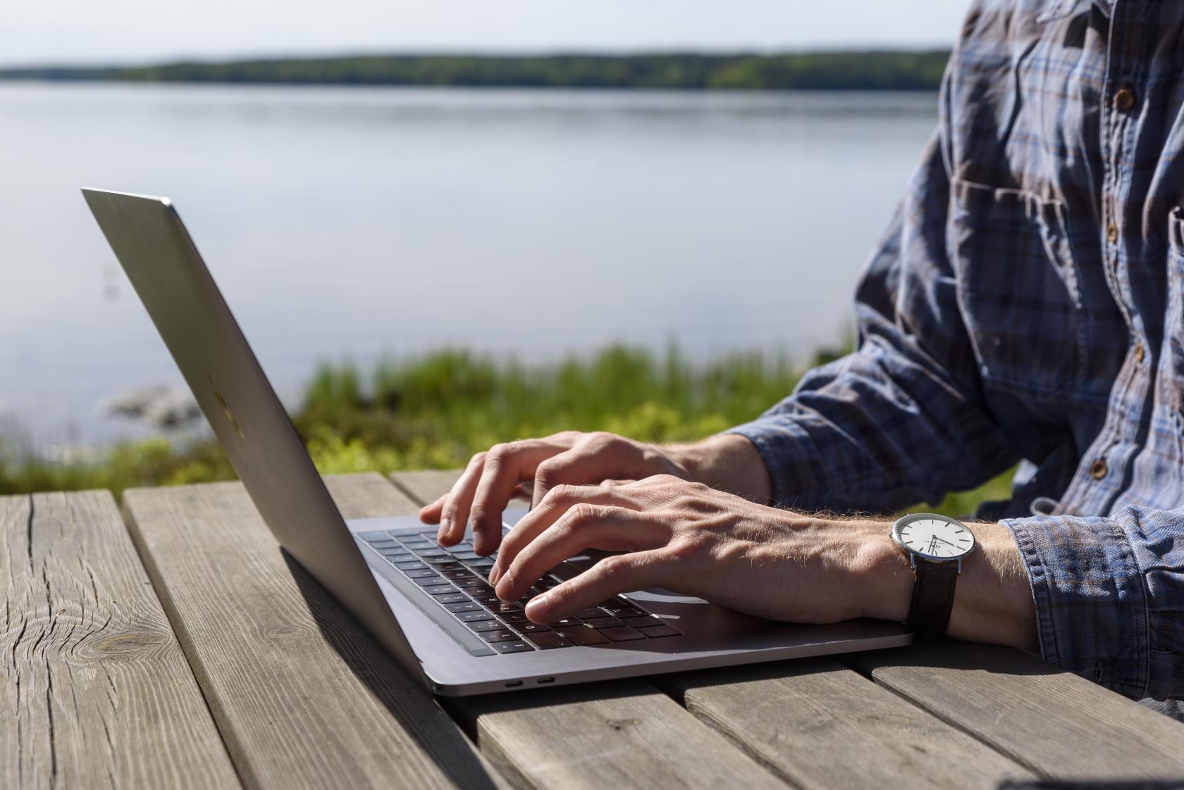 En person sitter vid ett träbord vid en sjö och jobbar på sin laptop.