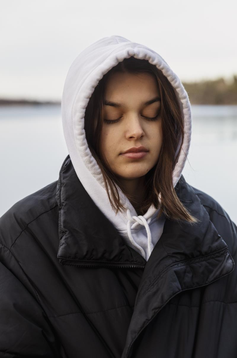 Ung kvinna iklädd hoodie och täckjacka. Hon tittar ner och ser allvarlig ut.