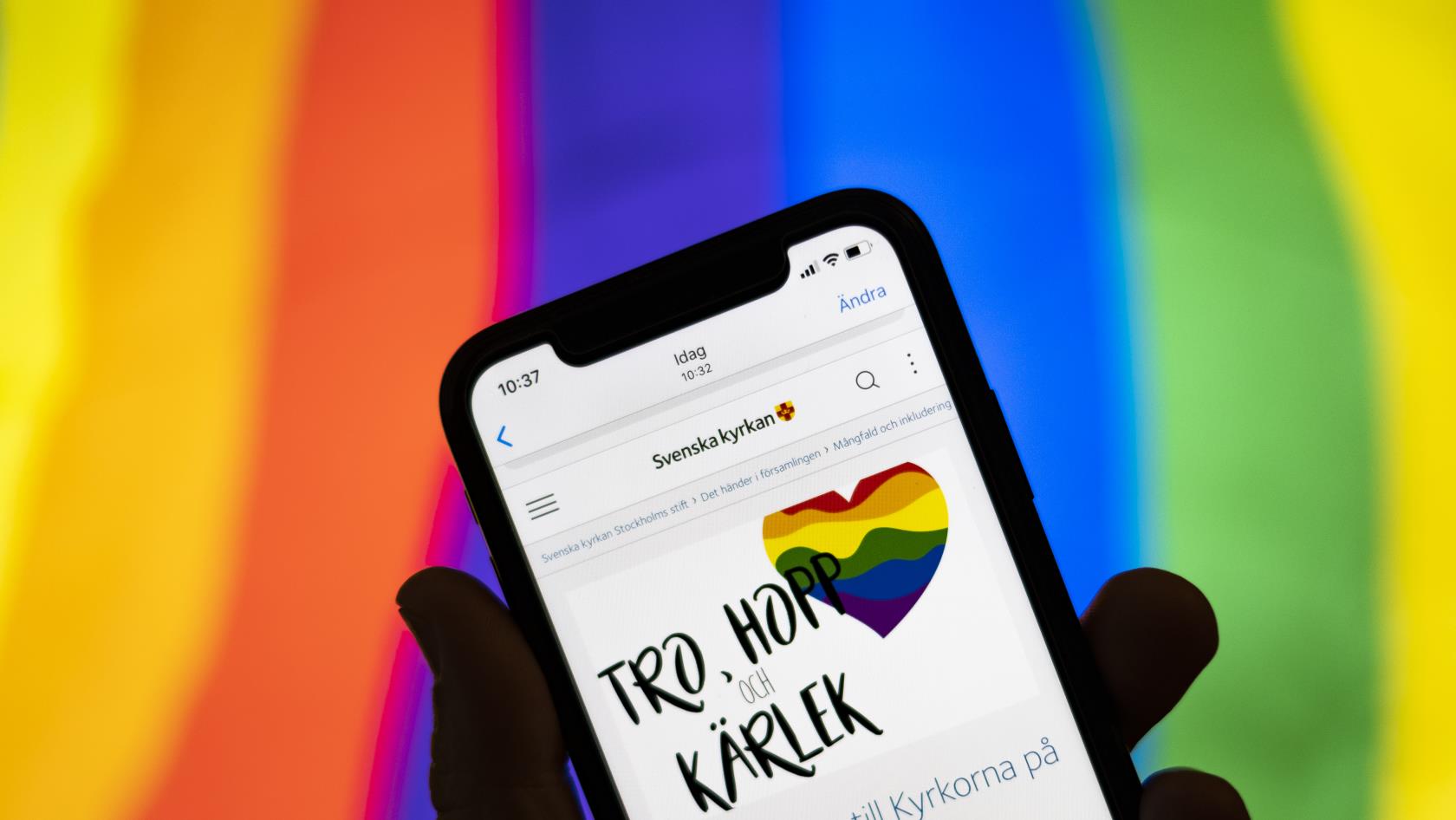 Någon håller en mobiltelefon där skärmen visar Svenska kyrkans information om Pride. I bakgrunden syns regnbågsfärgerna.