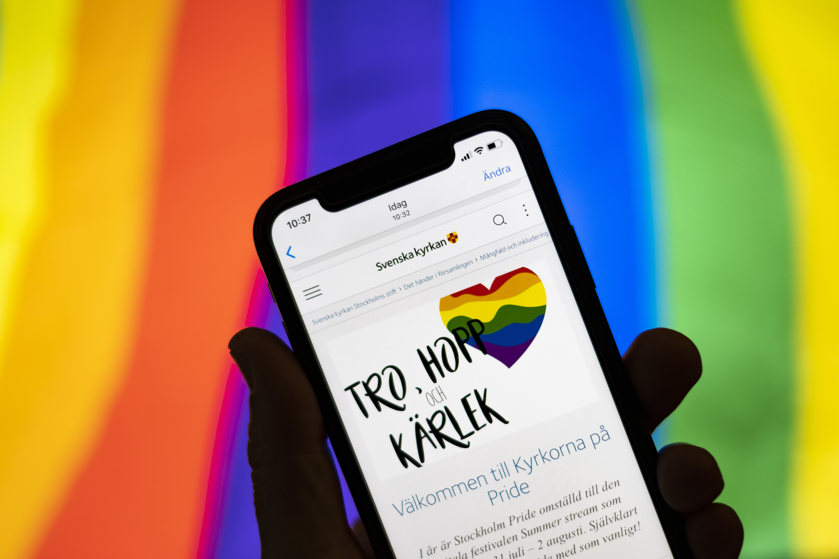 Någon håller en mobiltelefon där skärmen visar Svenska kyrkans information om Pride. I bakgrunden syns regnbågsfärgerna.