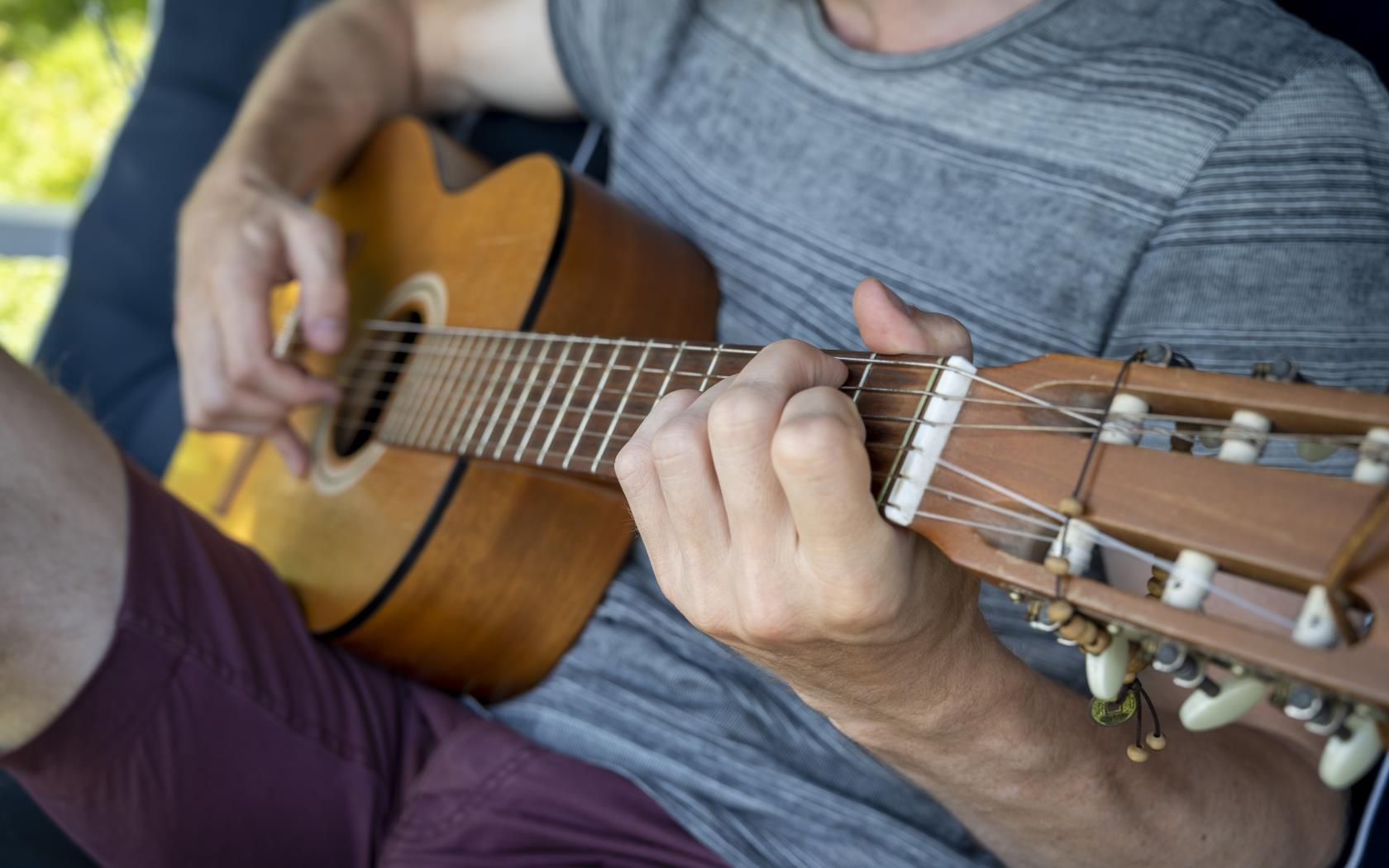 En man sitter utomhus och spelar gitarr. Solen skiner.