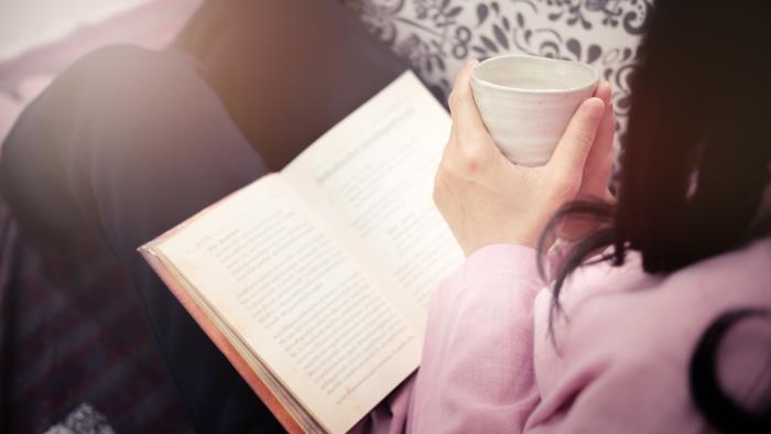En kvinna sitter och läser med en bok i knät och en kopp i händerna.