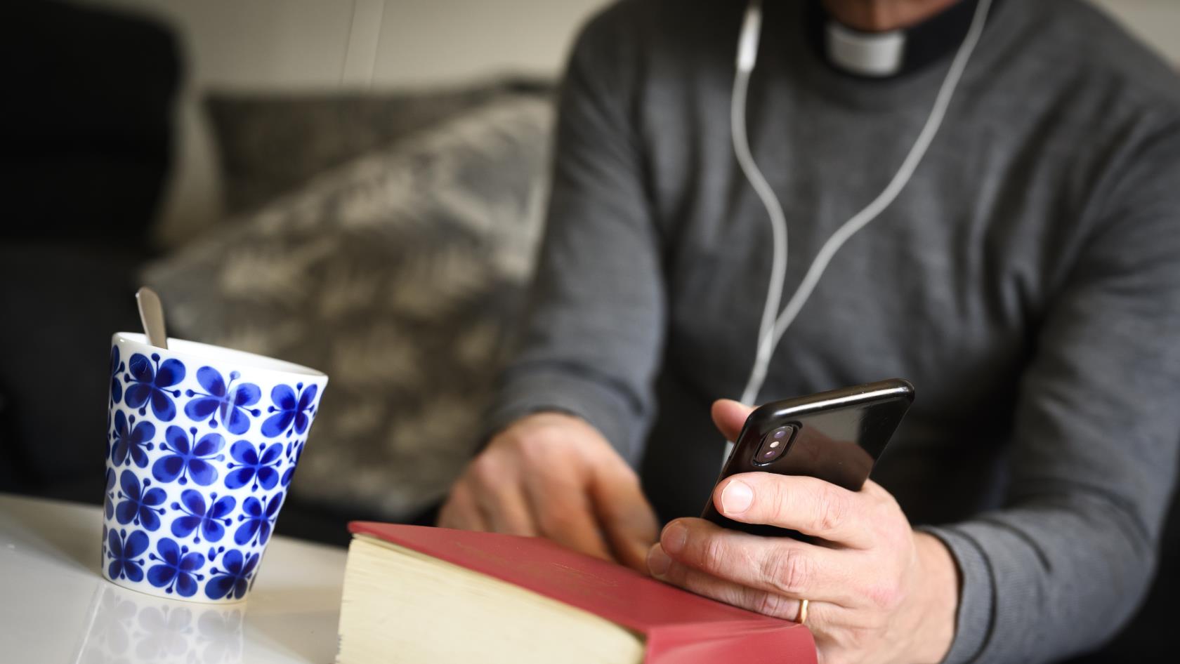 En präst sitter med hörlurar och pratar i mobiltelefon. En psalmbok ligger på bordet framför.