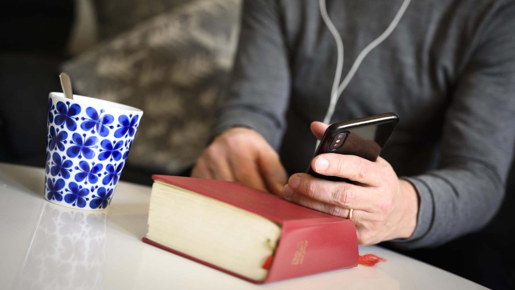 En präst sitter med hörlurar och pratar i mobiltelefon. En psalmbok ligger på bordet framför.