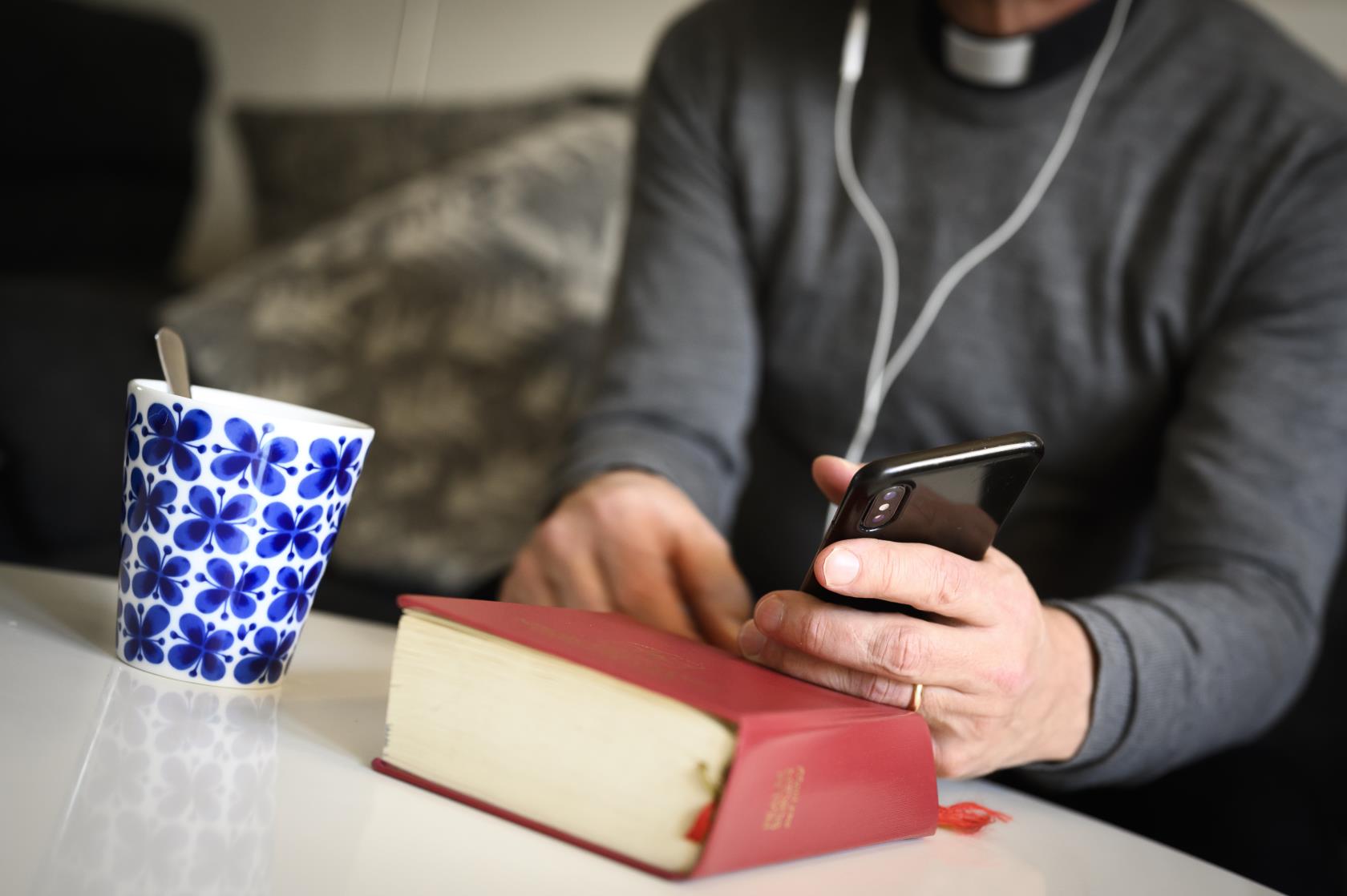 En präst sitter med hörlurar och pratar i mobiltelefon. En psalmbok ligger på bordet framför.