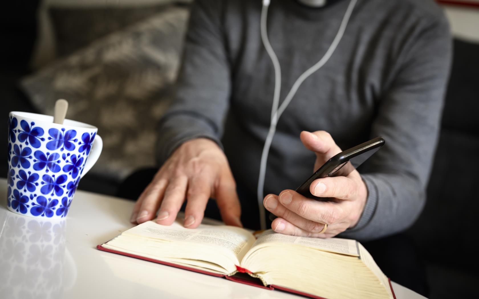 En präst sitter med hörlurar och pratar i mobiltelefon med bibeln uppslagen framför sig.