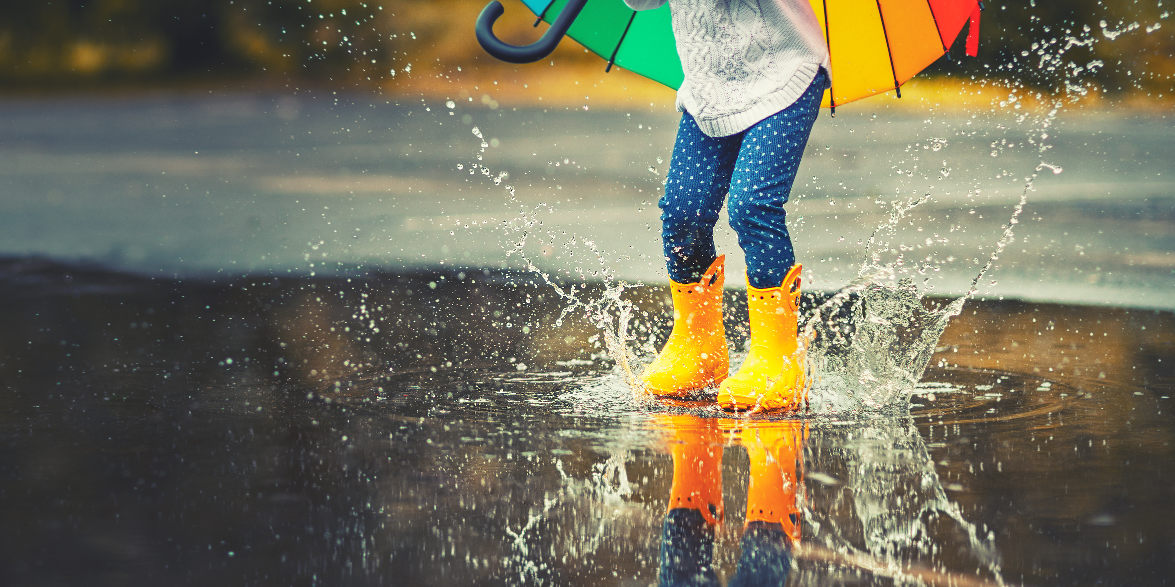 Ett barn med ett färgglatt paraply i handen och gula gummistövlar hoppar i en vattenpöl.