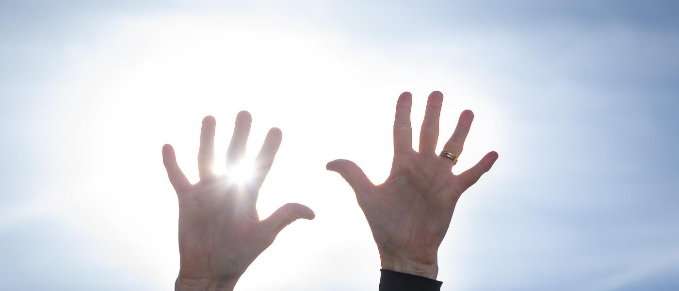 Två händer uppsträckta mot en blå himmel. Solen lysen mellan fingrarna.