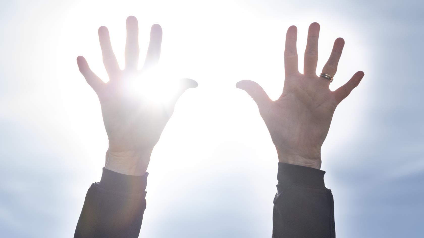 Två händer uppsträckta mot en blå himmel. Solen lysen mellan fingrarna.