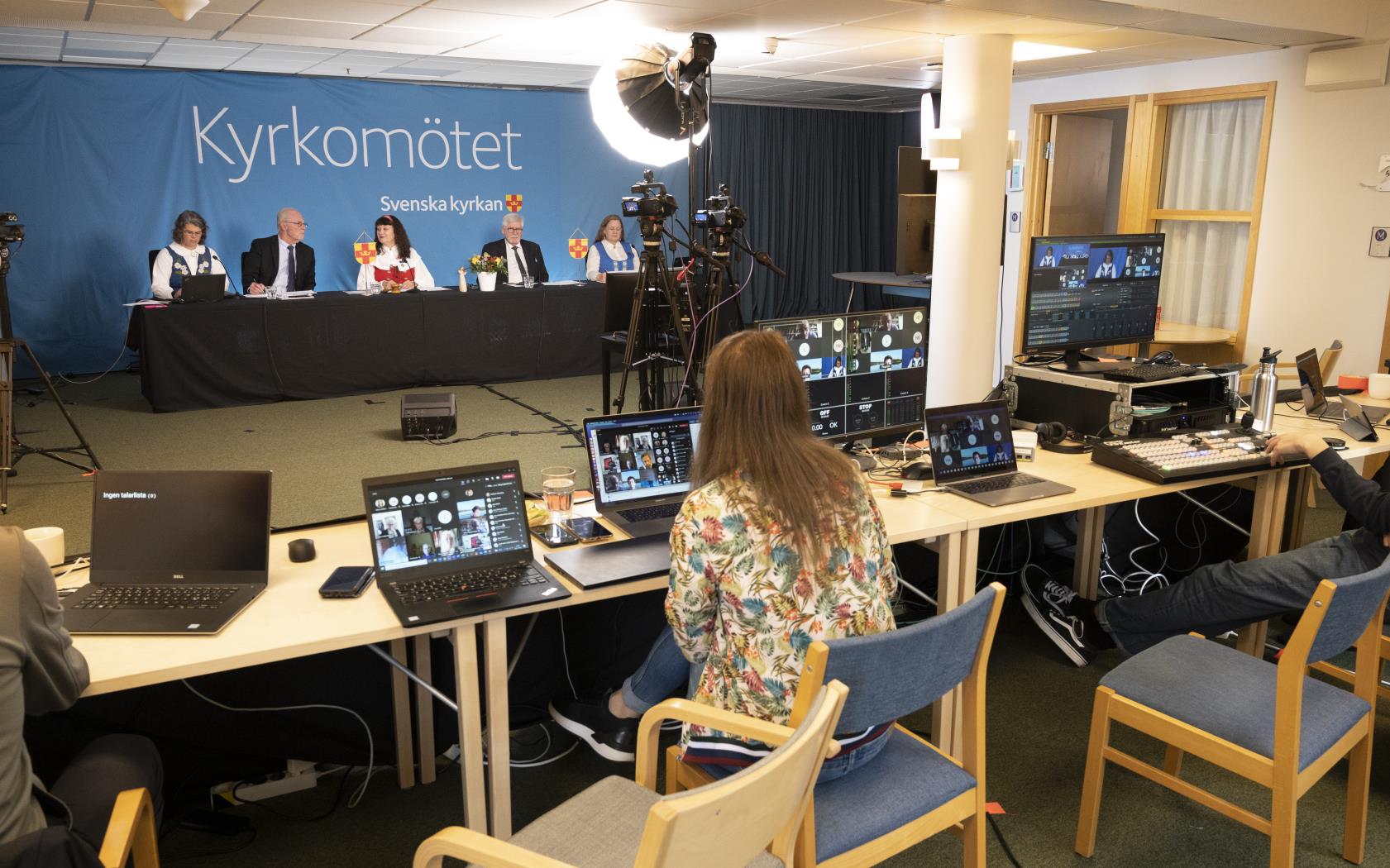 Kyrkomötets mötesledare sitter på en scen med filmkameror riktade mot sig. Bildtekniker med datorskärmar i förgrunden.