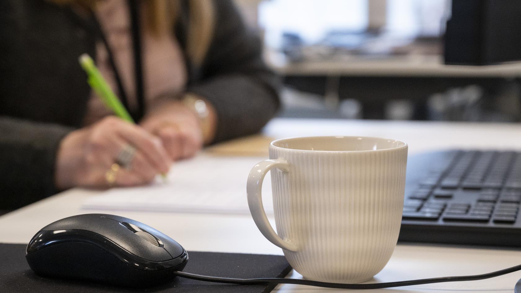 En kaffekopp står bredvid en datormus på ett skrivbord. En kvinna som antecknar syns suddigt i bakgrunden.